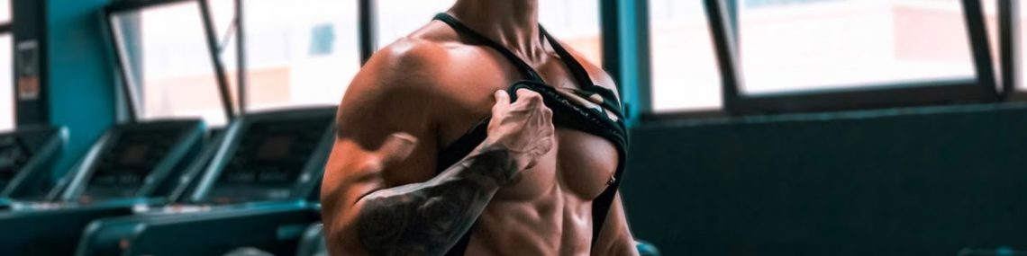Muscular man showing abs and tattoos in a gym setting.