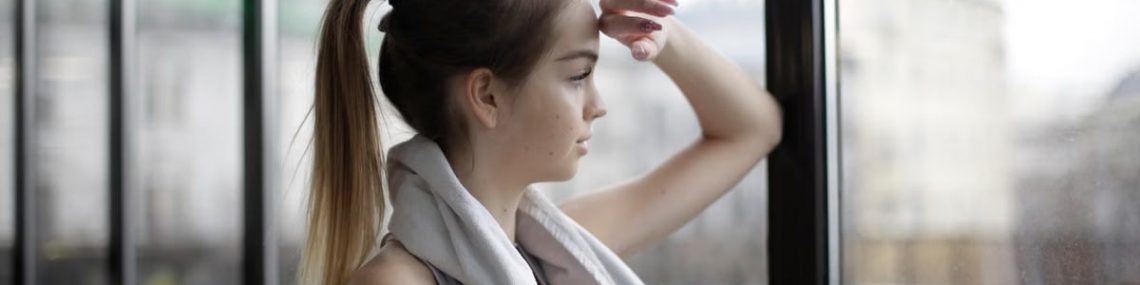Side view of thoughtful young female athlete in sportswear with towel around neck standing near window with elbow on glass and looking away pensively while resting after training in modern gym